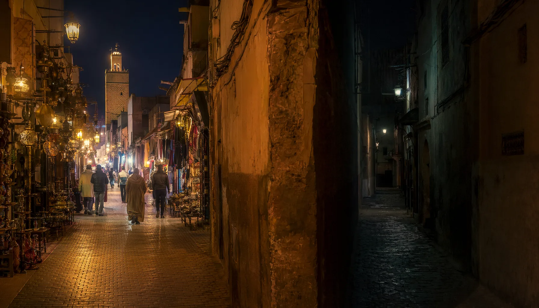 Ruelle de la médina de Marrakech la nuit, axe principal éclairé par des lanternes en contraste avec un derb sombre en arrière-plan
